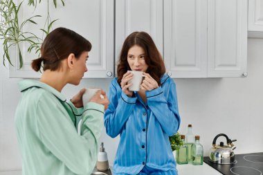 Pijama giyen genç lezbiyen bir çift, mutfakta sakin bir sabahın tadını çıkarıyor, sıcak bir içecek paylaşıyorlar..