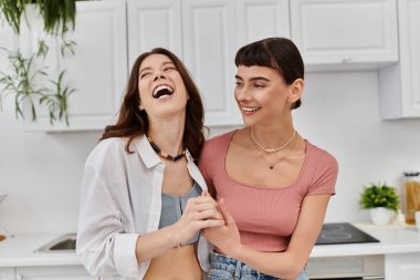 Two women laugh and hold hands in a cozy kitchen, enjoying a morning filled with love and happiness. clipart