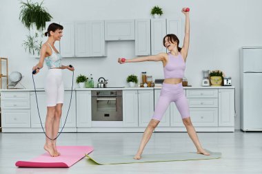 Spor giyimli iki genç kadın modern apartman mutfağında birlikte çalışıyor. Biri atlama halatı, diğeri ağırlık kaldırıyor..