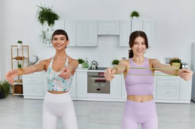 Spor giyimli, gülümseyen, evinde direniş bandıyla egzersiz yapan iki genç kadın..