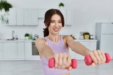 Sporcu genç bir kadın gülümserken pembe halterler takar. Beyaz dolaplı, modern bir mutfakta egzersiz yapıyor..