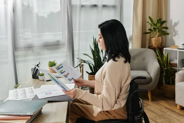 Stock image A woman in a wheelchair sits at a desk, carefully examining data charts.