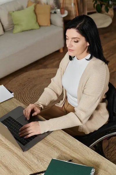 stock image A woman in a wheelchair sits at a desk, working on a laptop.