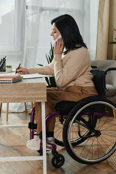 stock image A woman in a wheelchair sits at her desk, taking notes on a call.