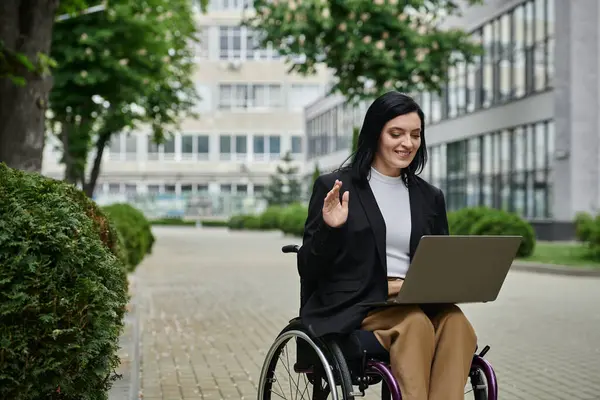 Stock image A woman in a wheelchair sits outside, smiling and waving during a video call on her laptop.