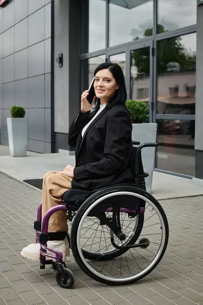 Tekerlekli sandalyedeki bir kadın modern bir binanın önünde telefonla konuşurken gülümsüyor..