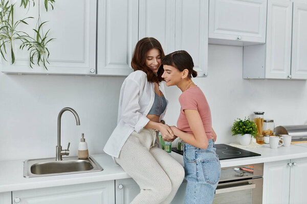 Two young women embrace and whisper in a bright kitchen, their love and joy evident in their smiles.