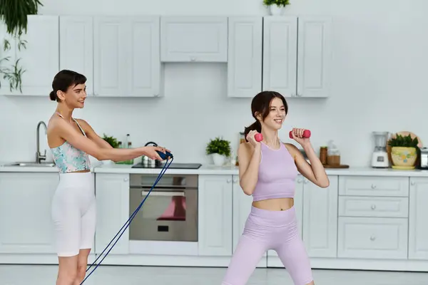 stock image Two young women in sportswear exercise in their modern kitchen. One woman is using a jump rope while the other lifts weights.