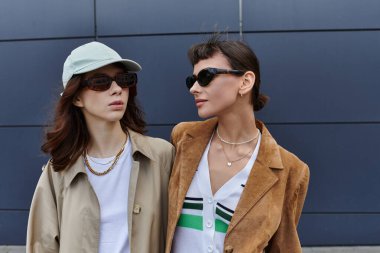 Şık giyinmiş iki genç kadın modern bir binanın önünde yan yana dururlar..