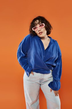 Mavi gömlekli ve güneş gözlüklü bir kadın turuncu arka planda duruyor..