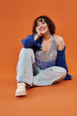 Bir kadın sıcak turuncu bir zemine oturuyor, parlak bir bluz ve kot pantolon giyiyor..