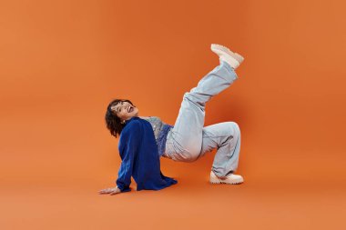 Mavi gömlek ve kot pantolon giyen bir kadın parlak turuncu arka planda poz verirken güler..