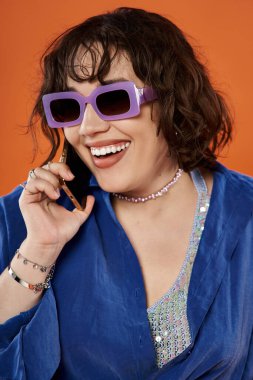 Mor güneş gözlüklü bir kadın telefonuyla konuşurken gülümsüyor, pullu mavi bir bluz giyiyor..