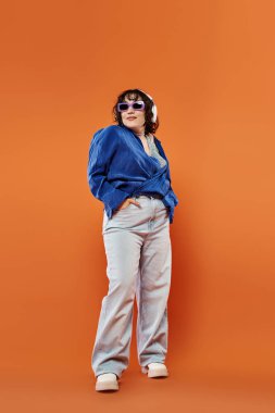Mavi gömlekli, beyaz pantolonlu ve güneş gözlüklü kadın turuncu arka planda güvenle duruyor.