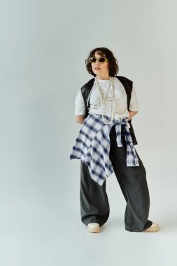 Ekose gömlekli ve siyah pantolonlu bir kadın güvenli bir şekilde beyaz bir stüdyoda duruyor..