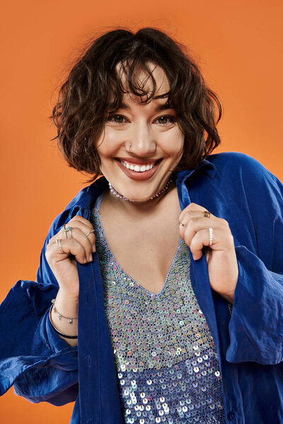 Женщина ярко улыбается в голубой рубашке, стоя на ярком оранжевом фоне.