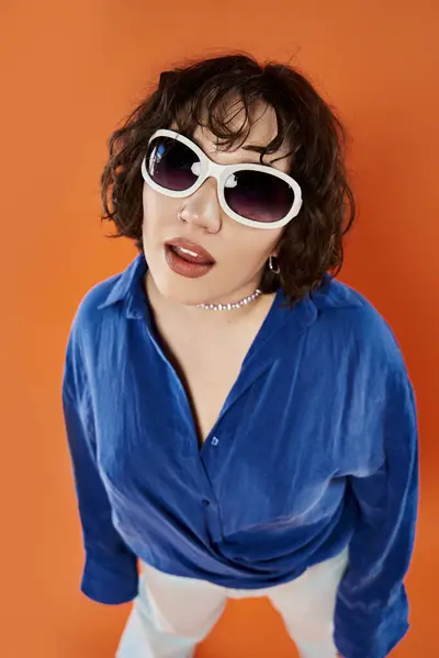 stock image A woman wearing sunglasses and a blue shirt poses against an orange background.