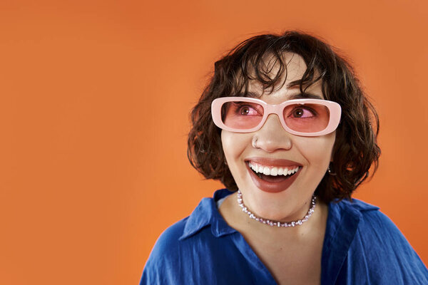 Женщина в солнечных очках ярко улыбается на ярком оранжевом фоне.