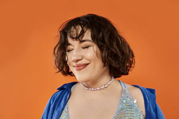 stock image Woman with brown hair smiling in blue jacket and sparkly top against orange background