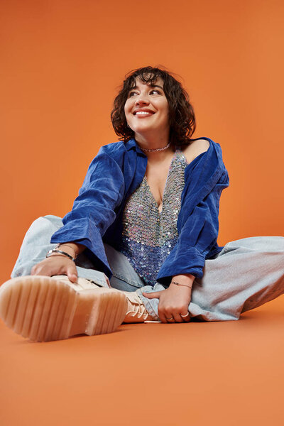 Женщина в голубой рубашке и блестящий топ сидит на оранжевом фоне, глядя радостно