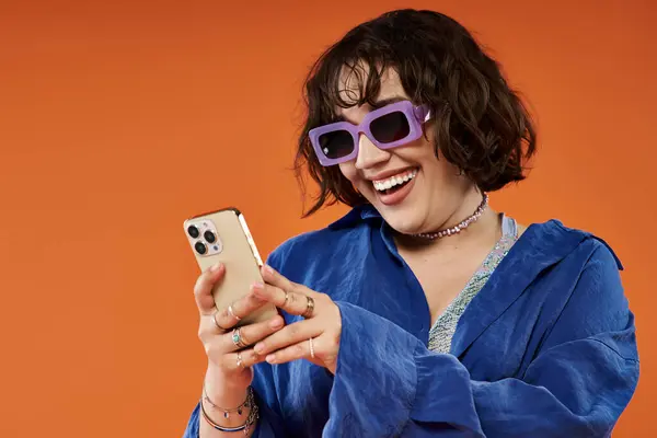 stock image A woman in sunglasses and a blue shirt laughs while reading her phone.