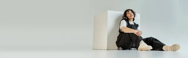 Stock image A woman sits thoughtfully on the floor, leaning against a white cube.