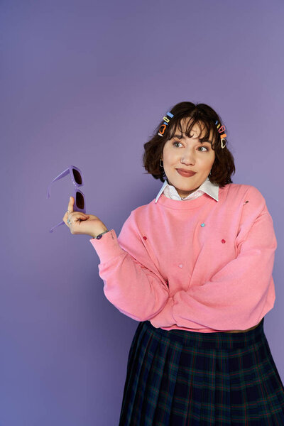 Женщина в розовом свитере и клетчатой юбке позирует на фиолетовом фоне.