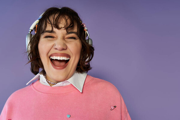 Женщина с короткими каштановыми волосами носит розовый свитер и широко смеется, наушники на шее.