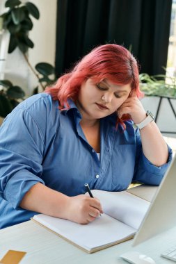 A plus size businesswoman with red hair and a blue shirt is focused on writing in her notebook. clipart