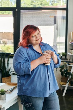 A plus size businesswoman with red hair stands near a window, holding a cup of coffee. clipart