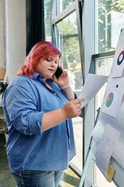 A plus size woman with bright red hair reviews charts while talking on the phone. clipart