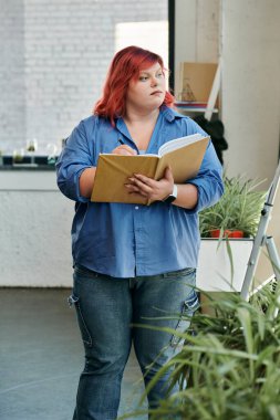 A plus size woman with bright red hair writes in a notebook, standing near a large plant. clipart