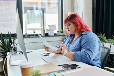 A plus size woman with bright red hair reviews documents at a desk in a contemporary office. clipart