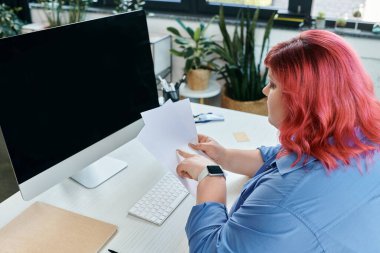 A plus size woman reviews papers at her desk with a computer screen in the background. clipart