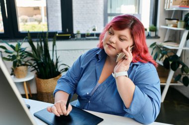 A plus size woman with bright pink hair takes a call while working on a drawing tablet at her desk. clipart