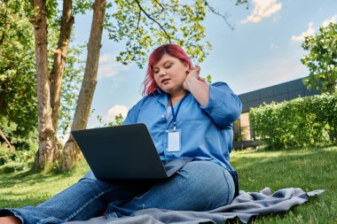 Pembe saçlı, iri yarı bir kadın parkta bir battaniyenin üzerinde oturur ve laptopuyla çalışır..