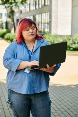 A plus size woman with pink hair uses her laptop while walking outside. clipart