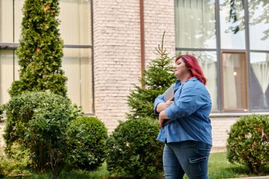 Kızıl saçlı artı cüsseli kadın dizüstü bilgisayarını tutarak güvenli bir şekilde parkta yürüyor.