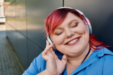 Parlak kızıl saçlı iri yarı bir kadın kulaklıkla müzik dinlerken gülümsüyor..