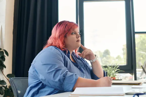 stock image A plus size businesswoman with pink hair, wearing a blue shirt, sits at a desk looking thoughtfully off to the side.