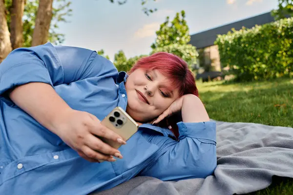 stock image A plus size woman with pink hair lies on a blanket outdoors, smiling as she uses her phone.