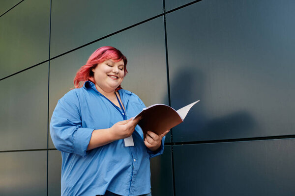 A plus size woman in a blue shirt stands against a steel wall, reading a pamphlet.