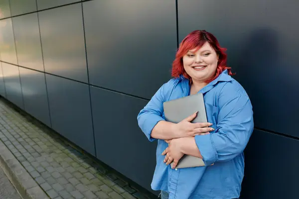 stock image A plus size woman with red hair smiles confidently while holding a laptop.