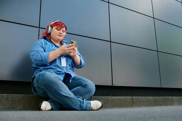 stock image A plus size woman sits on a curb in front of a modern building, lost in her phone.