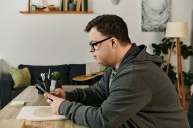 A young man with Down syndrome is focused on his phone while seated at his dining table. clipart