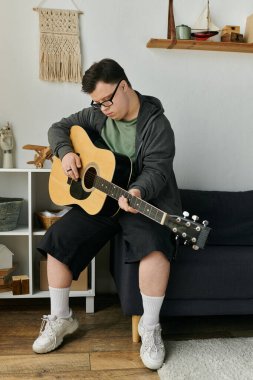 Down sendromlu genç bir adam, gitarını düşünceli bir şekilde yaşam alanında çalıyor..