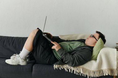 A man with Down syndrome relaxes on the couch, engaged with his laptop. clipart