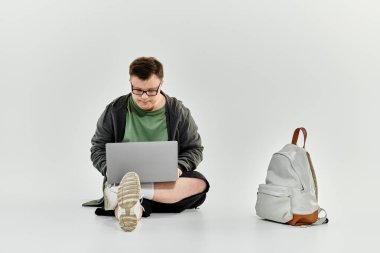 Down sendromlu genç bir adam yerde otururken dizüstü bilgisayarına odaklanıyor..