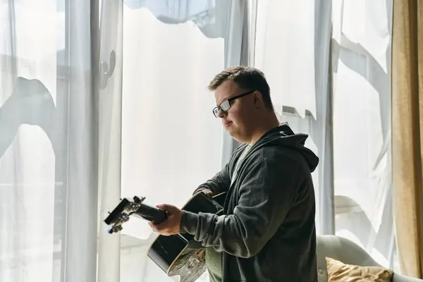 stock image A young man with Down syndrome strums a guitar, enjoying a sunny moment at home.