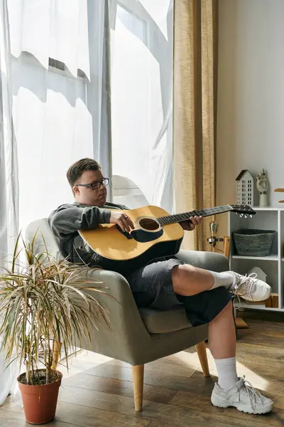 stock image A young man with Down syndrome plays guitar while relaxing in his comfortable chair at home.